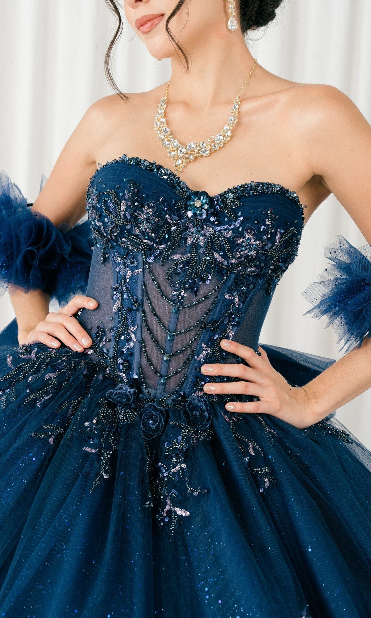 A woman wears the Quinceanera Dress 8380J by Cinderella Couture, a strapless navy blue ball gown with floral and bead embellishments, glitter tulle sleeves, a corset bodice, and a jeweled necklace.